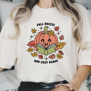 May include: A white t-shirt with a graphic of a smiling pumpkin reading a book surrounded by autumn leaves. The text "Fall Breeze and Cozy Reads" is printed below the pumpkin.