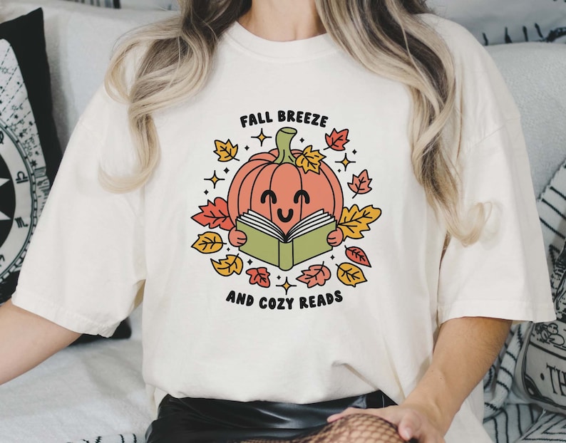 May include: A white t-shirt with a graphic of a smiling pumpkin reading a book surrounded by autumn leaves. The text "Fall Breeze and Cozy Reads" is printed below the pumpkin.
