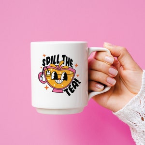 May include: A white ceramic mug featuring a cartoon graphic of a smiling orange teacup with a tea bag. The text "SPILL THE TEA!" is printed in bold black letters above and below the cup, accented by small orange star shapes on a white background.