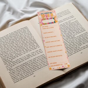 May include: A pink and orange bookmark with a floral design and the text "It's a great day to read". The bookmark has a section for writing down book details, including the date started, favorite character, and how the book made the reader feel.