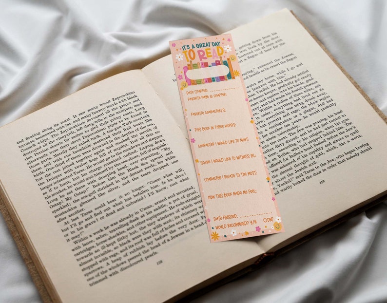 May include: A pink and orange bookmark with a floral design and the text "It's a great day to read". The bookmark has a section for writing down book details, including the date started, favorite character, and how the book made the reader feel.