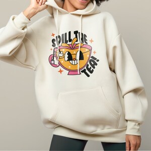 May include: A cream-colored hooded sweatshirt featuring a graphic of a smiling, anthropomorphic teacup with a tea bag. The text "SPILL THE TEA!" is printed in black above and to the side of the cup, surrounded by small orange star icons.