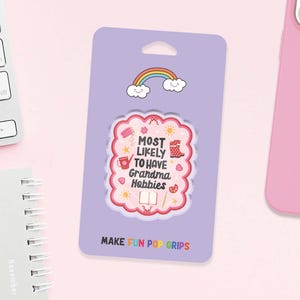 Może przedstawiać: Jasnofioletowy PopGrip z r&oacute;żowym i białym wzorem z napisem "MOST LIKELY TO HAVE Grandma Hobbies". PopGrip ma u g&oacute;ry tęczę i chmury i znajduje się na jasnofioletowym tle. Tekst "MAKE FUN POP GRIPS" znajduje się na dole.