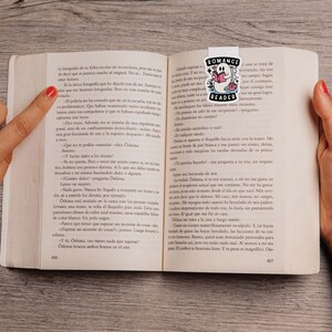 Op de afbeelding: Een close-up van een open boek met een bladwijzer met een spook dat een boek leest. Het boek is getiteld "Romance Reader". De tekst op de pagina luidt: "Que no te pod&iacute;as controlar -dice Oshima. -Y, Oshima, &iquest;tienes nada que superar? Oshima levanta ambos brazos en el aire."