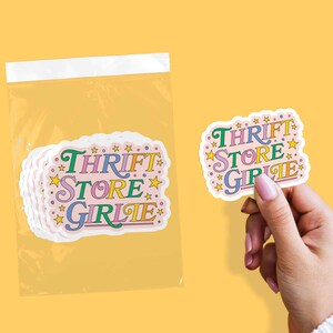 May include: A stack of stickers in a clear plastic bag and one sticker held by a hand. The stickers have a pink background with the words "THRIFT STORE GIRLIE" in colorful text and star accents.