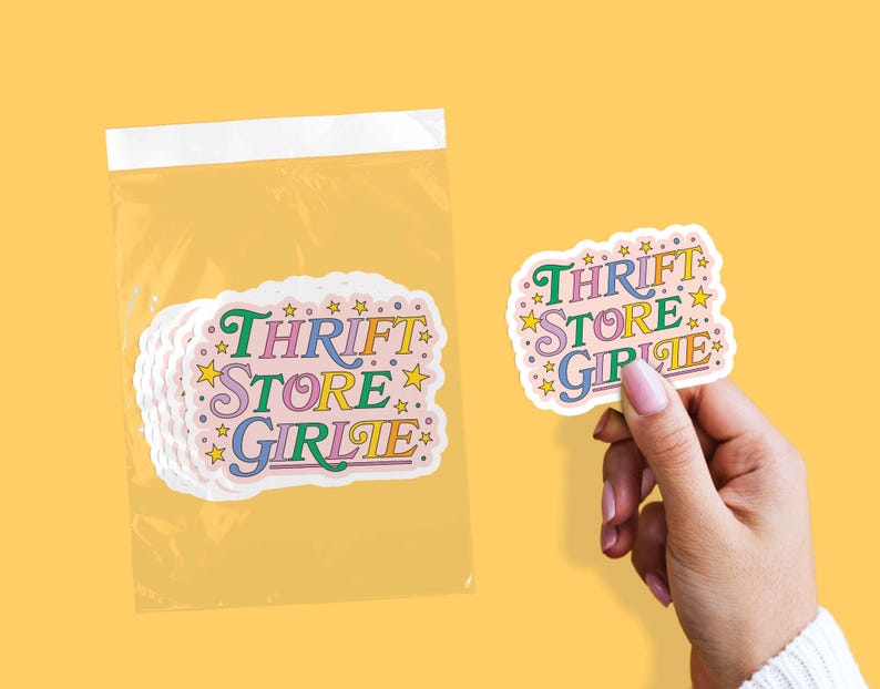 May include: A stack of stickers in a clear plastic bag and one sticker held by a hand. The stickers have a pink background with the words "THRIFT STORE GIRLIE" in colorful text and star accents.