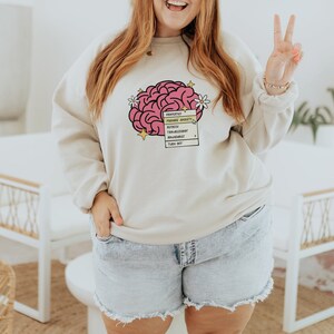 May include: A beige sweatshirt with a pink brain graphic and a checklist that reads "Prioritize", "Manage Anxiety", "Reflect", "Transcend", "Flourish", "Bountiful", "Think Big".