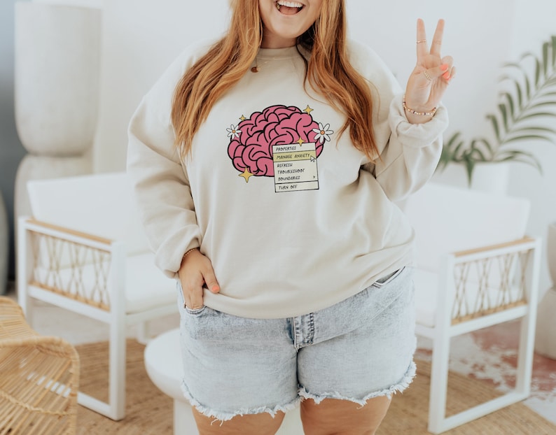 May include: A beige sweatshirt with a pink brain graphic and a checklist that reads "Prioritize", "Manage Anxiety", "Reflect", "Transcend", "Flourish", "Bountiful", "Think Big".