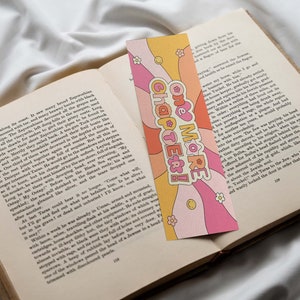 K&ouml;nnte beinhalten: Ein rosa und gelbes Lesezeichen im Retro-Design. Das Lesezeichen zeigt ein Herz mit dem Text "One More Chapter!" in einem wei&szlig;en Kreis. Das Lesezeichen ist mit Blumen und Schn&ouml;rkeln verziert.