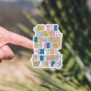 May include: A colorful sticker with the text "Be the reason someone believes in the goodness of people" in a retro font. The sticker has a white border and small flower designs.