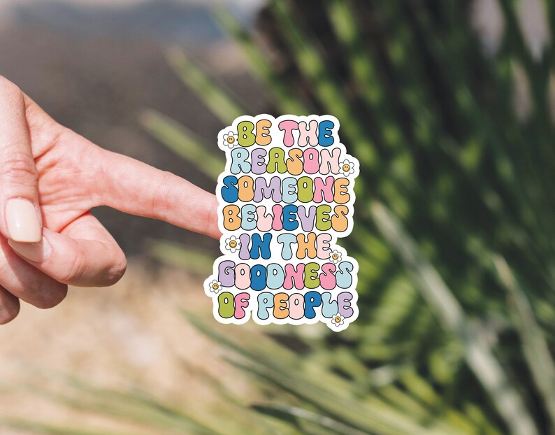 May include: A colorful sticker with the text "Be the reason someone believes in the goodness of people" in a retro font. The sticker has a white border and small flower designs.