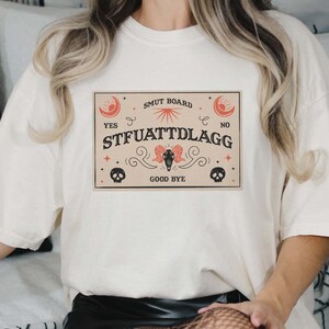 Puede incluir: Camiseta blanca con un gr&aacute;fico de una tabla Ouija de estilo vintage con el texto "SMUT BOARD", "YES", "STFUATTDLAGG", "NO" y "GOOD BYE".