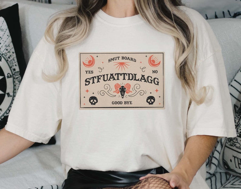 Puede incluir: Camiseta blanca con un gr&aacute;fico de una tabla Ouija de estilo vintage con el texto "SMUT BOARD", "YES", "STFUATTDLAGG", "NO" y "GOOD BYE".