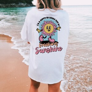 Op de afbeelding: Wit T-shirt met een cartoon zon-afbeelding en de tekst "I'm a happy go lucky ray of freaking sunshine".