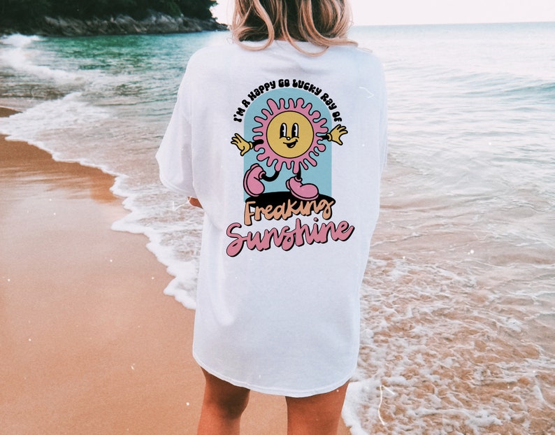Op de afbeelding: Wit T-shirt met een cartoon zon-afbeelding en de tekst "I'm a happy go lucky ray of freaking sunshine".
