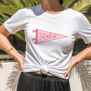 May include: White t-shirt with a pink pennant graphic that says "Tired AF".