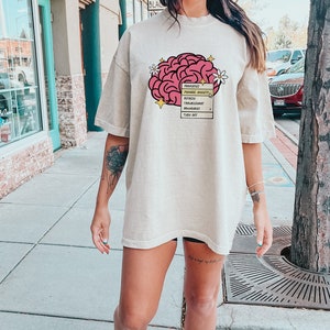 May include: An oversized beige t-shirt featuring a pink brain graphic and the text "Prioritize, Manage Anxiety, Reflect, Trust Your Instincts, Boundaries, Turn Off", a flower graphic, and a yellow star graphic.