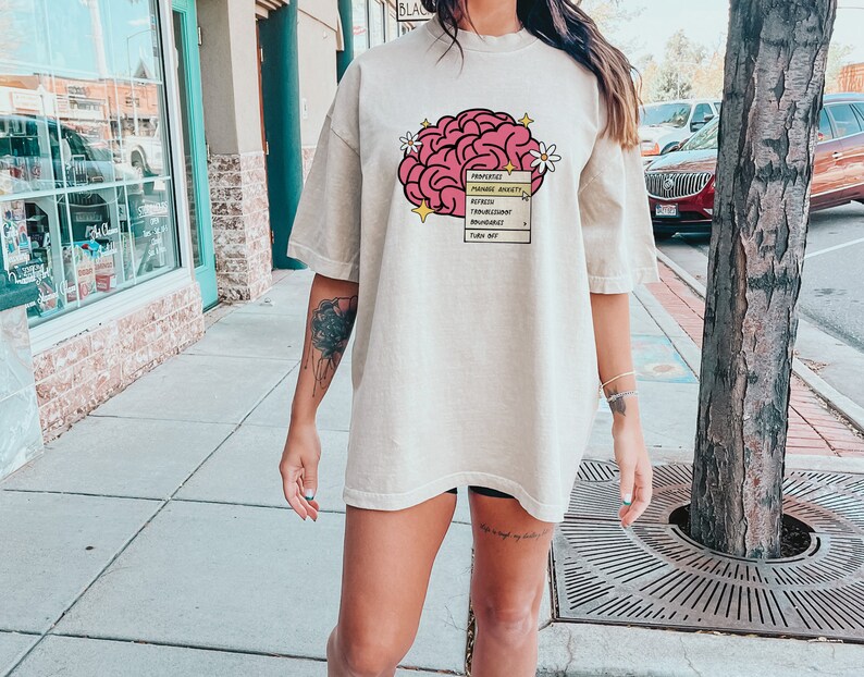 May include: An oversized beige t-shirt featuring a pink brain graphic and the text "Prioritize, Manage Anxiety, Reflect, Trust Your Instincts, Boundaries, Turn Off", a flower graphic, and a yellow star graphic.