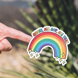 K&ouml;nnte beinhalten: Ein Regenbogen-Aufkleber mit dem Text "Queer, All Day, All Year" in wei&szlig;er Umrandung. Der Aufkleber hat auf jeder Seite des Regenbogens zwei kleine Sterne.
