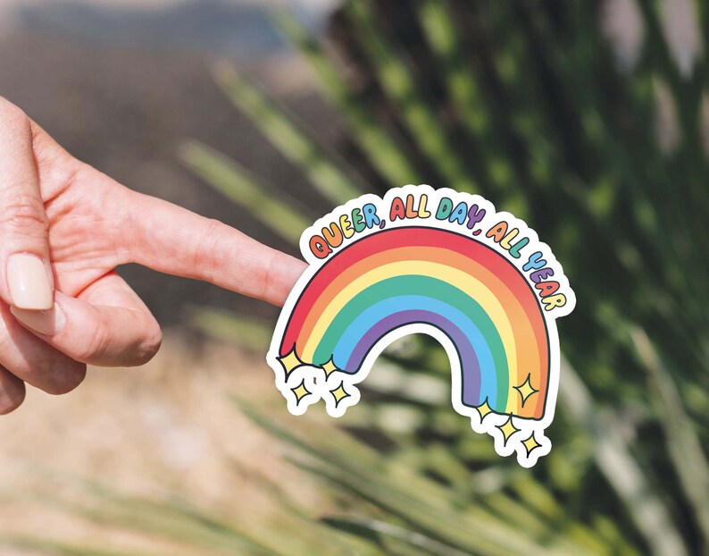 K&ouml;nnte beinhalten: Ein Regenbogen-Aufkleber mit dem Text "Queer, All Day, All Year" in wei&szlig;er Umrandung. Der Aufkleber hat auf jeder Seite des Regenbogens zwei kleine Sterne.