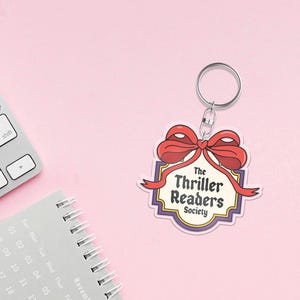 May include: A keyring featuring a design with a red bow and the words "The Thriller Readers Society". The design is set against a white and purple background. The background is a light pink colour.