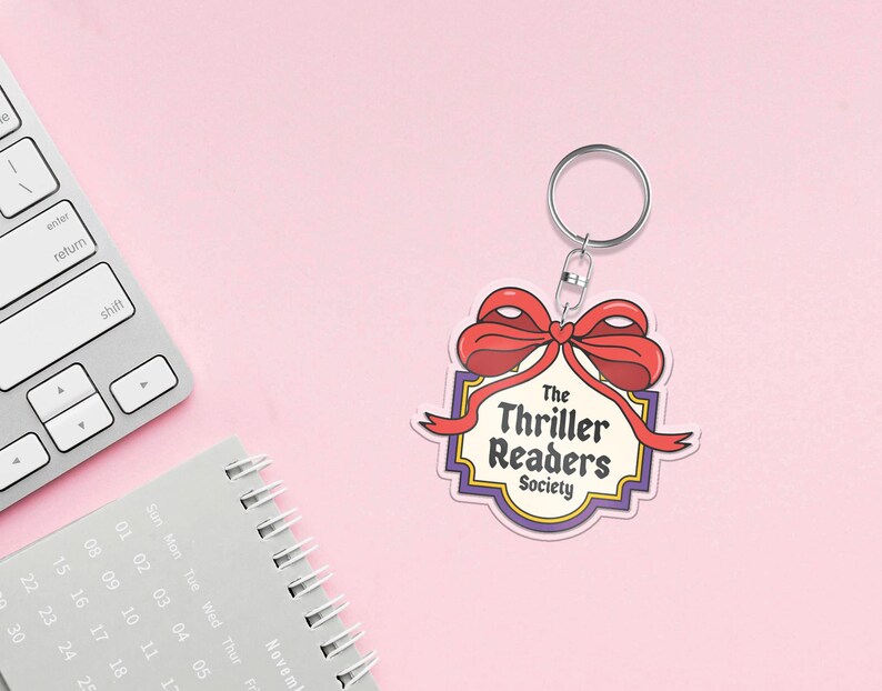 May include: A keyring featuring a design with a red bow and the words "The Thriller Readers Society". The design is set against a white and purple background. The background is a light pink colour.