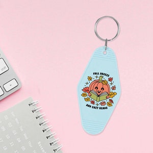 May include: A light blue keychain shaped like a motel key with a smiling pumpkin reading a book. The text on the keychain reads "Fall Breeze and Cozy Reads".