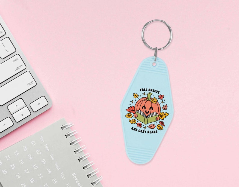 May include: A light blue keychain shaped like a motel key with a smiling pumpkin reading a book. The text on the keychain reads "Fall Breeze and Cozy Reads".