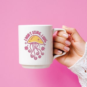 K&ouml;nnte beinhalten: Eine wei&szlig;e Keramiktasse mit einer rosa und gelben Illustration einer Qualle und dem Text "Ich steche nicht, ich umarme."