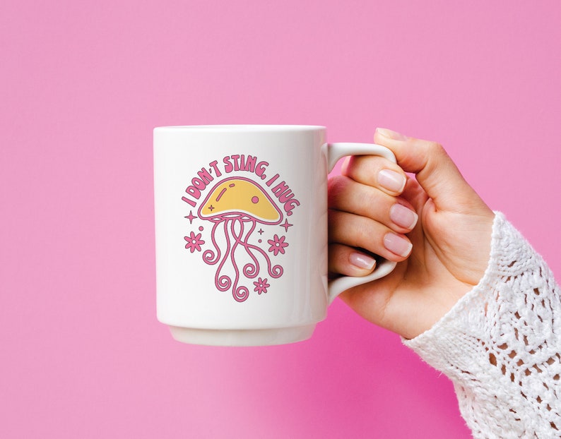 K&ouml;nnte beinhalten: Eine wei&szlig;e Keramiktasse mit einer rosa und gelben Illustration einer Qualle und dem Text "Ich steche nicht, ich umarme."
