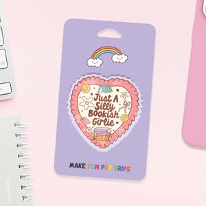 May include: A purple Pop Grip package with a heart-shaped design that reads "Just A Silly Bookish Girlie." The design includes a rainbow, clouds, flowers, and a strawberry. The text "MAKE FUN POP GRIPS" is at the bottom.