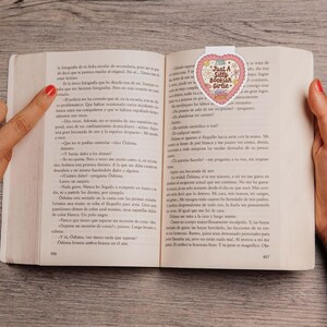 May include: A book is open to a page with text, held by two hands. A heart-shaped bookmark with the text "Just A Silly Bookish Girlie" is inserted in the book. The bookmark has a pink and white design.