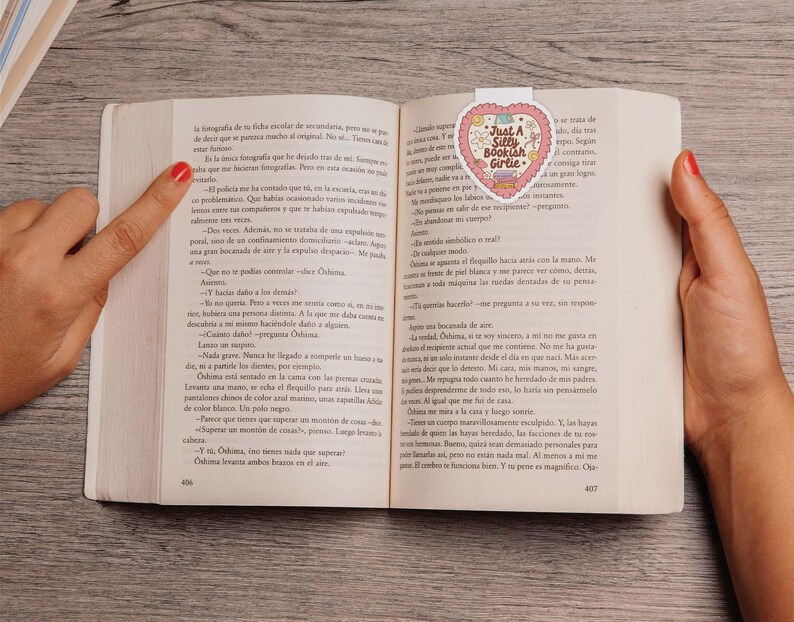 May include: A book is open to a page with text, held by two hands. A heart-shaped bookmark with the text "Just A Silly Bookish Girlie" is inserted in the book. The bookmark has a pink and white design.