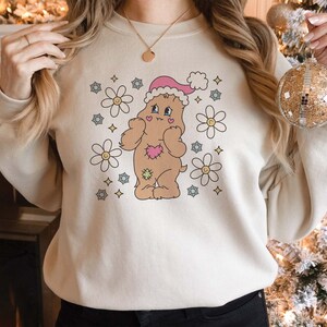 May include: A cream-coloured sweatshirt featuring a cartoon bear wearing a Santa hat and surrounded by flowers. The bear is brown with a pink heart on its chest.