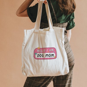 Peut inclure: Un sac fourre-tout en toile blanche avec un autocollant rose et blanc qui dit "Hello I am Dog Mom".