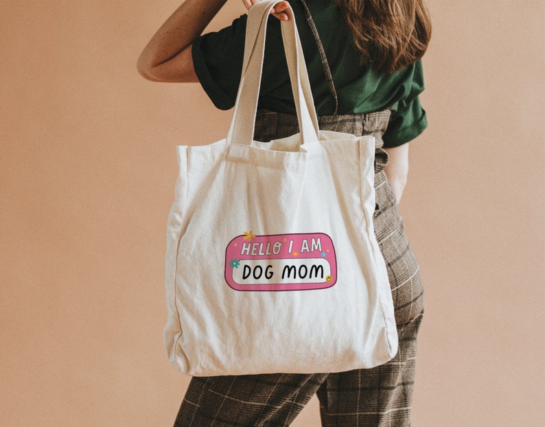 Peut inclure: Un sac fourre-tout en toile blanche avec un autocollant rose et blanc qui dit "Hello I am Dog Mom".