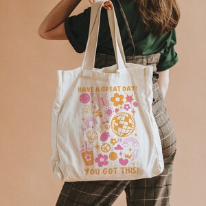 Puede incluir: Una bolsa de lona blanca con un dise&ntilde;o rosa y amarillo. La bolsa tiene el texto "Have a great day! You got this!" y presenta una variedad de divertidas ilustraciones de estilo retro, incluyendo flores, mariposas, una bola de discoteca y un signo de paz.