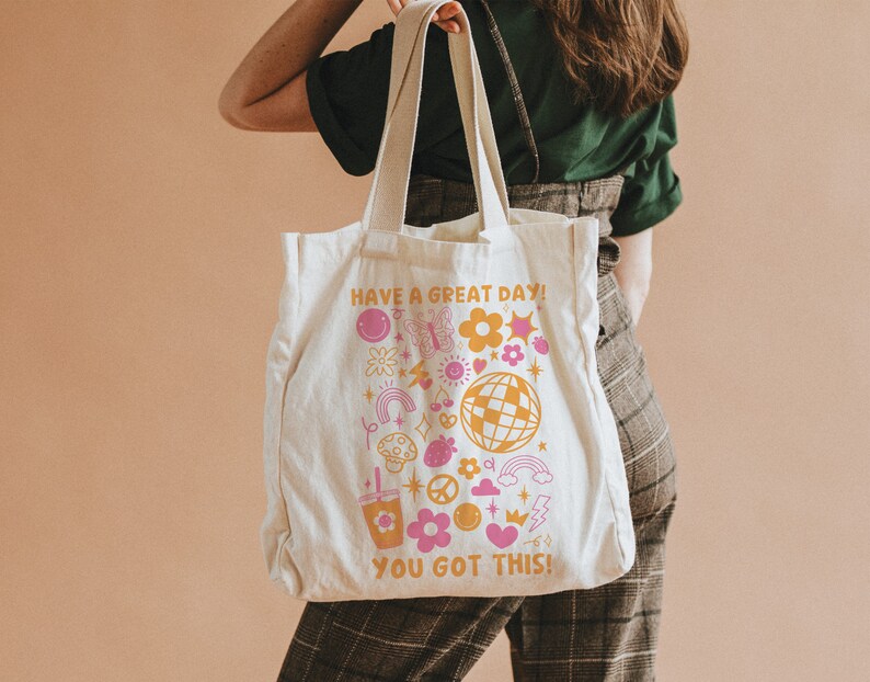 Puede incluir: Una bolsa de lona blanca con un dise&ntilde;o rosa y amarillo. La bolsa tiene el texto "Have a great day! You got this!" y presenta una variedad de divertidas ilustraciones de estilo retro, incluyendo flores, mariposas, una bola de discoteca y un signo de paz.