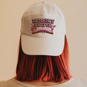 May include: White baseball cap with a pink and black graphic of a book with flames and the text "Romance Reader".