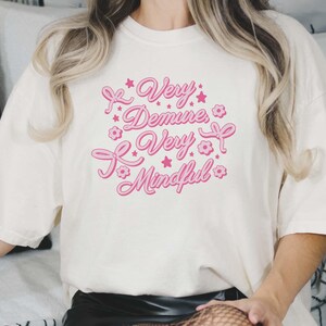 May include: White t-shirt with pink text that reads "Very Demure, Very Mindful" with pink stars and flowers around the text.