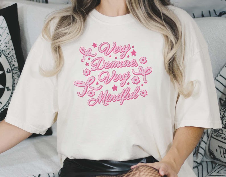 May include: White t-shirt with pink text that reads "Very Demure, Very Mindful" with pink stars and flowers around the text.