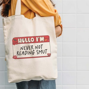 May include: A white canvas tote bag with a red and white name tag design that says "Hello I'm Never Not Reading Smut".