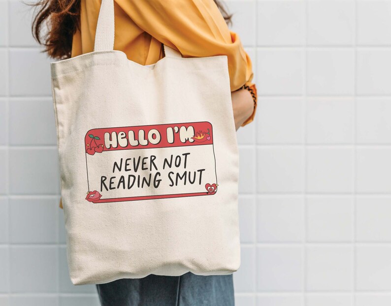 May include: A white canvas tote bag with a red and white name tag design that says "Hello I'm Never Not Reading Smut".