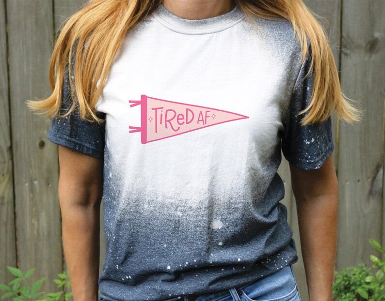 May include: A white t-shirt with a black and white ombre effect. The shirt has a pink pennant with the text "TIRED AF" printed on it.