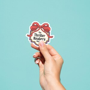 May include: A sticker featuring a red bow and the words "The Thriller Readers Society" within a decorative border. The sticker is held against a light blue backdrop.