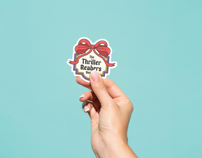 May include: A sticker featuring a red bow and the words "The Thriller Readers Society" within a decorative border. The sticker is held against a light blue backdrop.