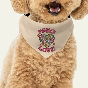 May include: A beige dog bandana with a pink heart design. The heart is filled with paw prints in various colors. The text "PAWS LOVE" is printed in pink letters on the bandana.