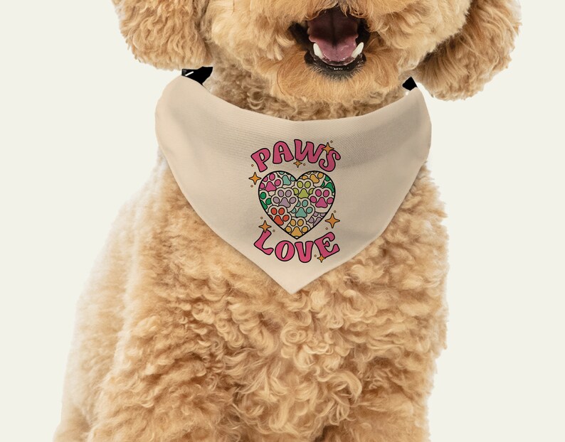 May include: A beige dog bandana with a pink heart design. The heart is filled with paw prints in various colors. The text "PAWS LOVE" is printed in pink letters on the bandana.