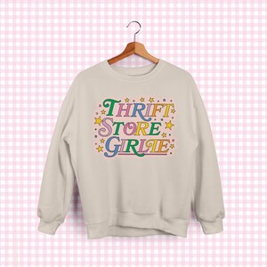 May include: A cream-colored sweatshirt with the text "Thrift Store Girlie" in colorful, retro-style lettering. The design includes stars and a playful font, creating a vintage aesthetic. The sweatshirt is hanging on a wooden hanger.