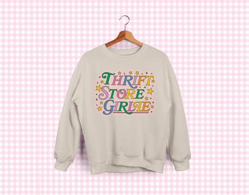 May include: A cream-colored sweatshirt with the text "Thrift Store Girlie" in colorful, retro-style lettering. The design includes stars and a playful font, creating a vintage aesthetic. The sweatshirt is hanging on a wooden hanger.